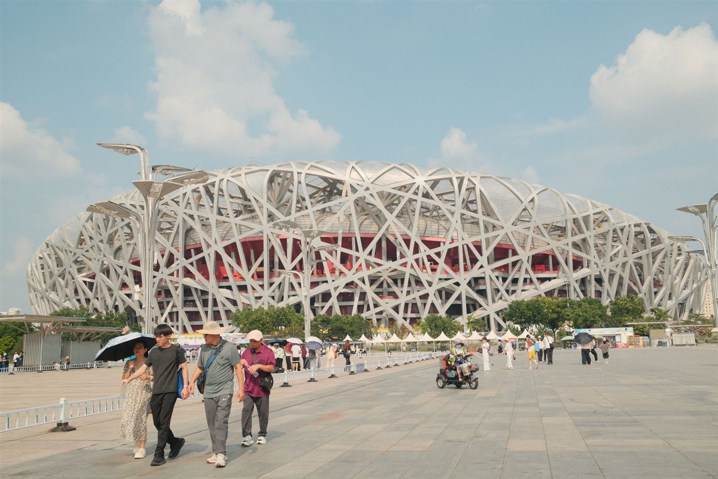 The steel lattice of the Bird’s Nest stadium under a blue sky, people walking across the plaza