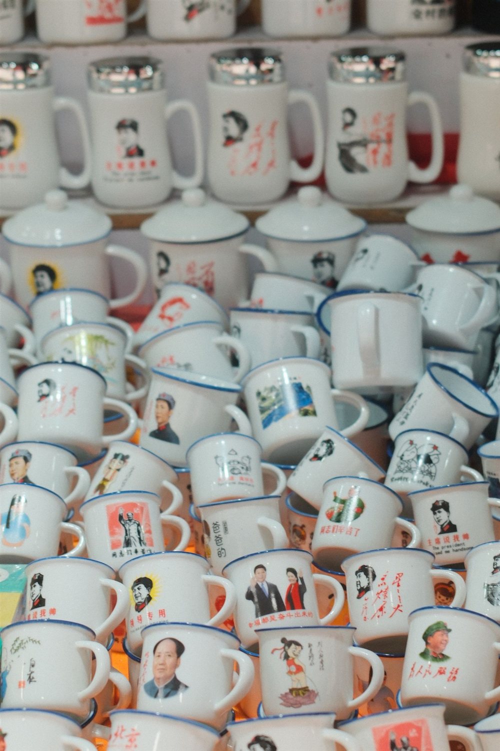 Rows of white enamel souvenir mugs with blue rims and printed portraits and slogans