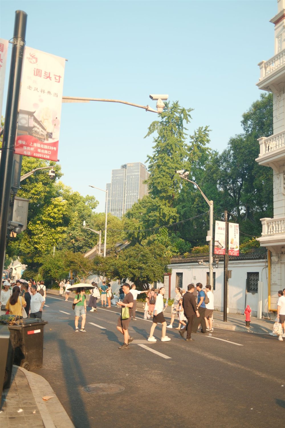 People crossing a sunlit street lined with trees and red banners